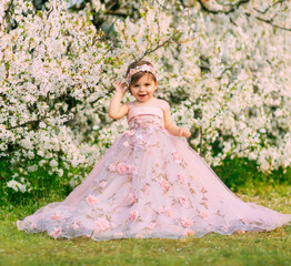 Obraz na płótnie Canvas Young happy little girl fairy princess. Happy child face is smiling. Luxurious fluffy long dress for children, pink outfit. White flowers background trees spring nature. Fashion model, one year old.