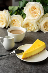 A piece of lemon cheesecake and coffee with milk on the table next to a bouquet of roses.