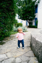 Fototapeta na wymiar Little girl stands on paving slabs near bushes