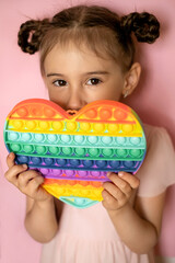 A girl with pigtails holds a heart-shaped popit toy in her hand and covers her face. Cute portrait of a child.  Beautiful girl on a pink background. Pop it toy. 
