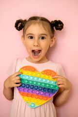 The girl with pigtails bulged her eyes in surprise, holding a popit toy in the shape of a heart in her hand and closing one eye. The child's mouth is open in admiration. Emotional portrait of a kid. 