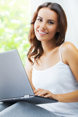 Portrait image of beautiful happy smiling brunette woman using laptop, at home, sitting against window.