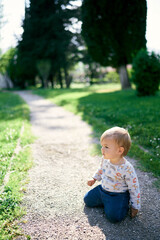 Toddler sits on his knees on the path in the green park. Side view