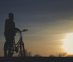 dziewczyna stojąca z rowerem na tle zachodu słońca - obrazy, fototapety, plakaty