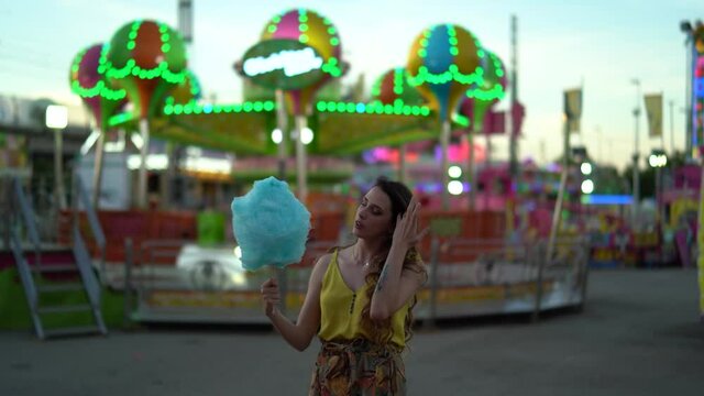 Chica joven con pelo largo tomando algod&oacute;n de az&uacute;car y pas&aacute;ndolo bien en zona de atracciones de una feria