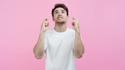 Young man in white t-shirt showing crossed fingers and looking up isolated on pink