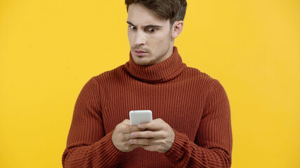 Worried man in sweater using smartphone isolated on yellow