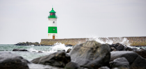 A white and green striped lighthouse at the end of a pier. In front of it are waves crashing...