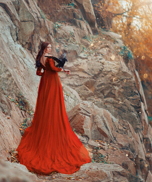 Fantasy woman with black dark raven bird on hand. Queen witch in red long vintage fashion dress, back rear view. Backdrop autumn nature, tree orange leaves. Fashion model posing, girl mystic princess