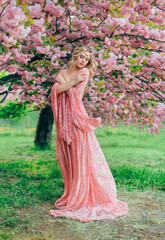 Fantasy happy woman princess walks in spring blooming garden hugs herself by shoulders. Pink...