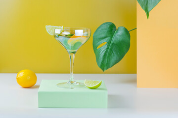 Cocktail in vintage champagne glass with mint leaves, lime and lemon on pastel color podiums. Summer cocktail with Monstera leaves on yellow and white background. Abstract concept. Part of set.
