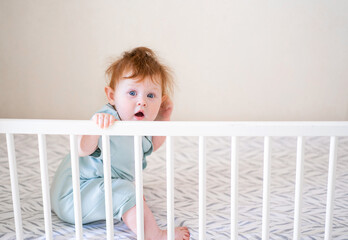 A baby with red hair is holding onto the side of the crib. Danger of the child falling out of bed.