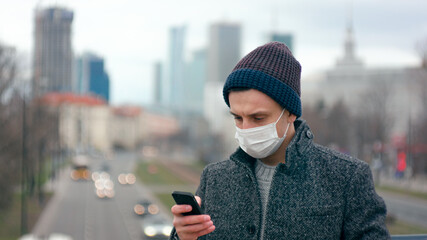 Casual Man in Surgical Face Mask Uses Mobile App on Smartphone Staying Outdside in Big City Downtown. COVID-19 Coronavirus Pandemic Outbreak or Smog. Background picture with Copy Space