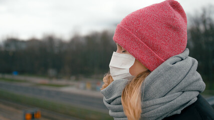 COVID-19 Coronavirus Pandemic or Smog Background. Portrait of Young Woman in Surgical Face Mask Looking at Big City Street in Winter. Picture with Copy Space