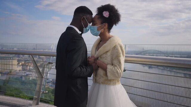 Positive loving African American couple in Covid-19 face masks rubbing noses holding hands standing on bridge. Happy bride and groom enjoying outdoor wedding on coronavirus pandemic