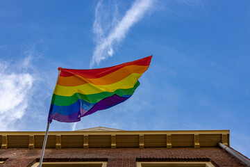 Dutch exterior facade of a home with rainbow colored LGBTQ+ flag contrasted strongly against a clear blue sky waving in a strong wind seen from below