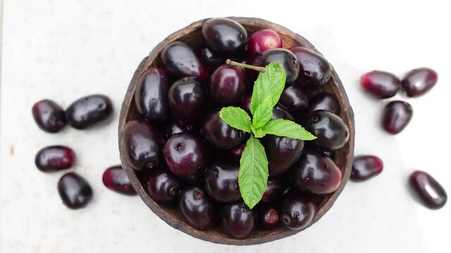 Dark pink-red ripe Syzygium cumini fruits. Dark black java plum in a wood bowl at isolated white background. Green mint leaf on top of some large java plums. 4k video.