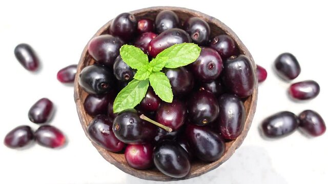 Dark pink-red ripe Syzygium cumini fruits. Dark black java plum in a wood bowl at isolated white background. Green mint leaf on top of some large java plums. 4k video.