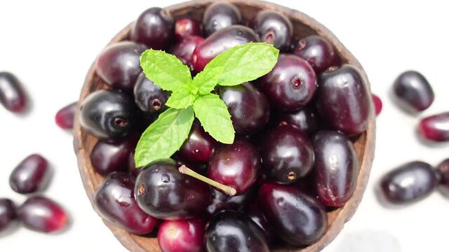 Dark pink-red ripe Syzygium cumini fruits. Dark black java plum in a wood bowl at isolated white background. Green mint leaf on top of some large java plums. 4k video.