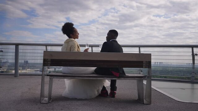 Wide shot of happy newlyweds toasting clinking glasses drinking alcohol sitting on bench outdoors. Positive loving African American bride and groom celebrating marriage on bridge