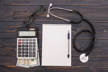 blank notepad with pen, stethoscope, calculator, dollars on a wooden background. Flat lay.