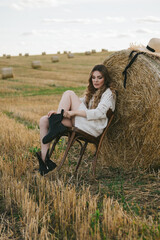 Naklejka na ściany i meble beautiful woman in a jacket and a hat with a chair by a haystack in the field.