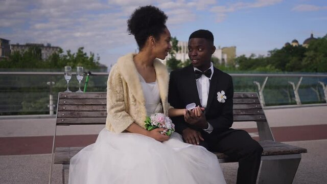 Positive bride and groom talking admiring luxurious wedding rings in box sitting on bench on sunny summer day. Happy African American couple getting married outdoors planning future