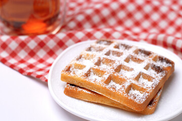 Homemade cakes - a recipe for Belgian waffles sprinkled with powdered sugar on a checkered tablecloth and served with black tea. Tasty breakfast at home.