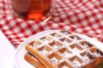 Homemade cakes - a recipe for Belgian waffles sprinkled with powdered sugar on a checkered tablecloth and served with black tea. Tasty breakfast at home.