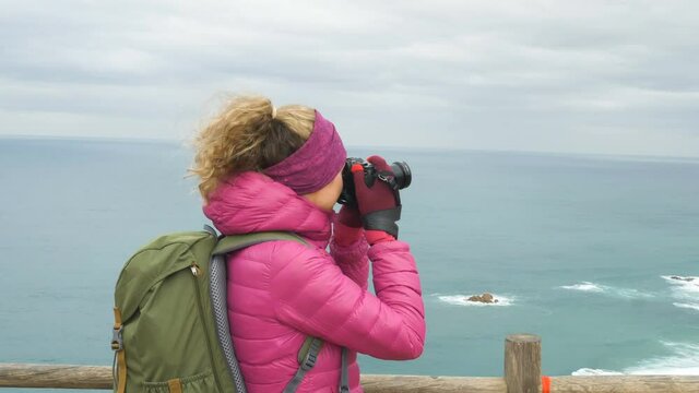 happy blonde girl tourist in pink jacket with backpack takes picture of beautiful ocean from viewing point on nasty day