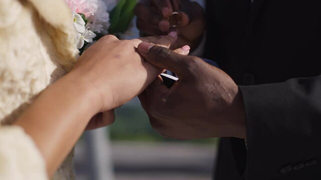 Close-up of African American male hand putting wedding ring on female finger outdoors. Unrecognizable loving couple getting married on sunny summer day. Love and marriage concept