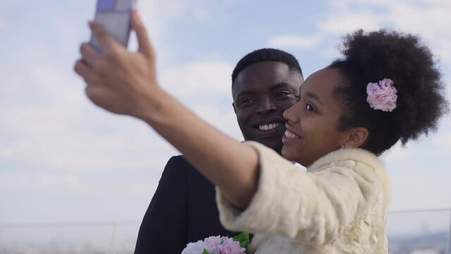 Cheerful smiling couple taking selfie on wedding ceremony on summer day outdoors. Happy young African American beautiful bride and handsome groom getting married photographing on smartphone