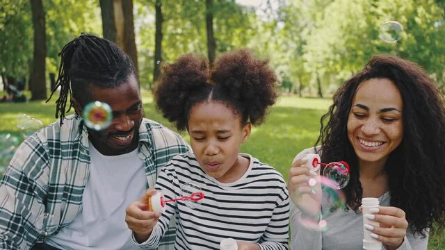 Close up of joyful African American famile with little kid girl blowing bubbles together, having fun on a picnic at city park, sitting on a blanket. Family time, vacations, leisure together
