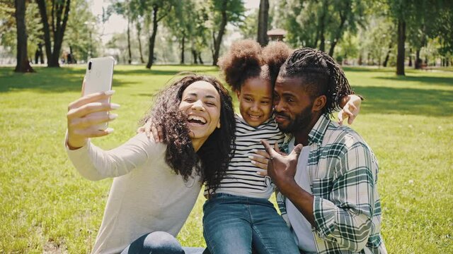 Lovely parents and their little daughter having fun in the nature, happy moments concept. African family takes a photo with a smartphone camera while walking in a summer park