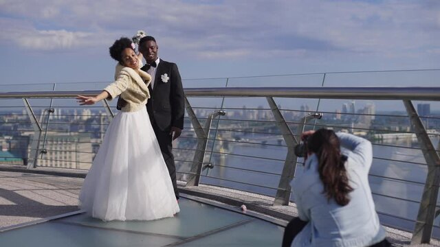 Wide shot of cheerful smiling African American bride and groom standing on bridge in sunlight posing for professional wedding photographer. Happy loving newlyweds photographing outdoors