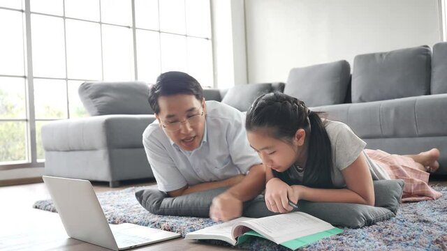 Asian family Father teaches daughter books by using laptop computer lying on the floor in the living room. Happy asian studying at home, study online