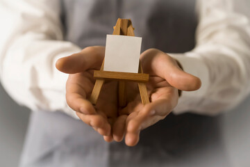 The male artist's hands are holding a miniature easel with a small piece of white paper on it. A...