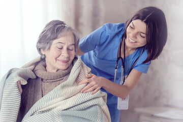 A young nurse takes care of an elderly 80-year-old woman at home, wraps a blanket around her. Happy retired woman and trust between doctor and patient. Medicine and healthcare.