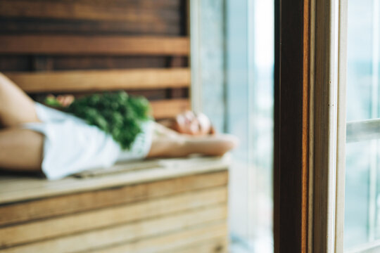 Blurred photo of Woman in white towel lies on wooden shelf in Finnish sauna with the green fresh broom, treat yourself