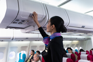 Young beautiful Asian airline cabin crew attendant closing overhead cabinet luggage compartment for...