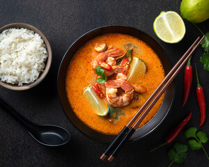 Tom Yum soup with shrimp in a ceramic black bowl with chopsticks on a dark background, top view