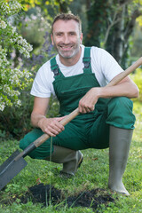 male gardener wearing overalls bent down holding a shovel