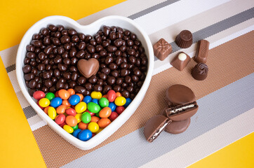 White heart-shaped bowl filled with brown and colorful chocolates, hazelnuts and almonds covered in chocolate
