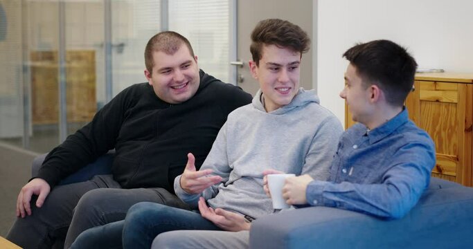 Team of young guys sitting on sofa in modern office emotionally discussing new project smiling laughing gesturing hands