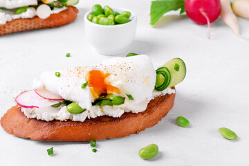 Poached egg on toasted bread with cream cheese, radish, cucumber and edamame beans on the white table. Breakfast sandwich