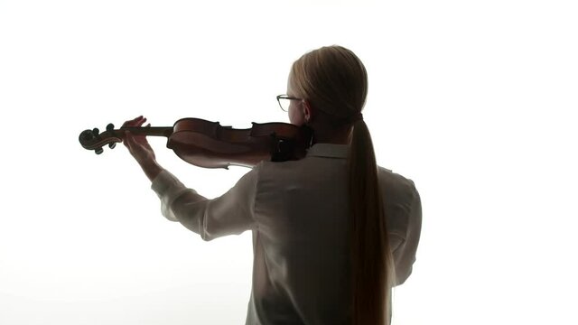 Woman blonde violinist in glasses and a blouse begins to play the violin, standing with back in a muffled light on white background of the studio.