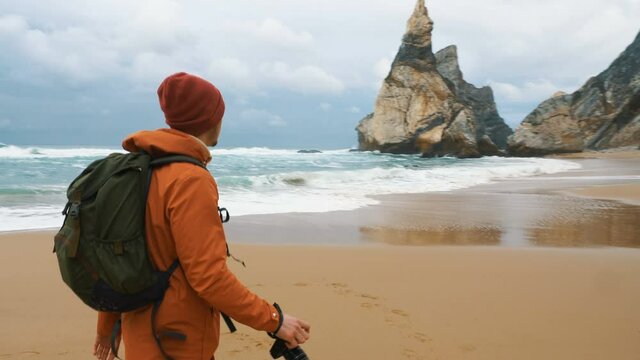 guy photographer with backpack takes picture of distant cliffs walking along empty ocean sand beach on nasty day