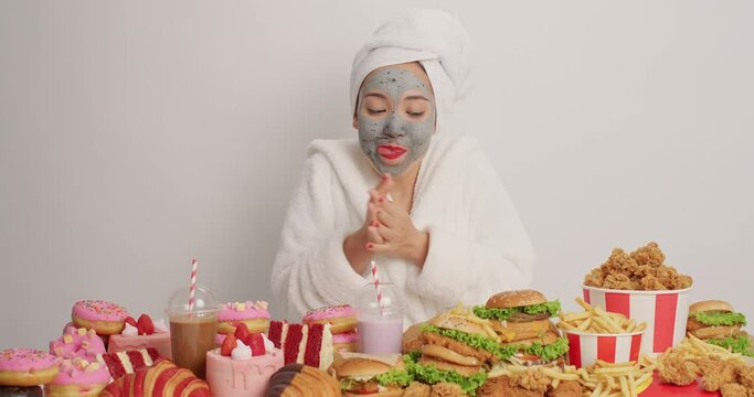 Mmm how delicious. Glad young woman wears bathrobe wrapped towel rubs hands starves to eat something tasty surroundedby junk food suffers from overeating habit isolated over white background