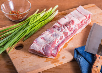 Cooking pork ribs. A large piece of raw pork belly with ribs on a cutting board with a butcher knife and a bunch of green onions.