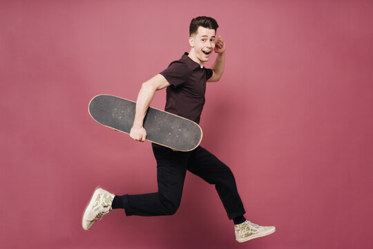 Portrait of young fashionable hipster man posing over pink background jumping. Caucasian male holding skateboard and smiling